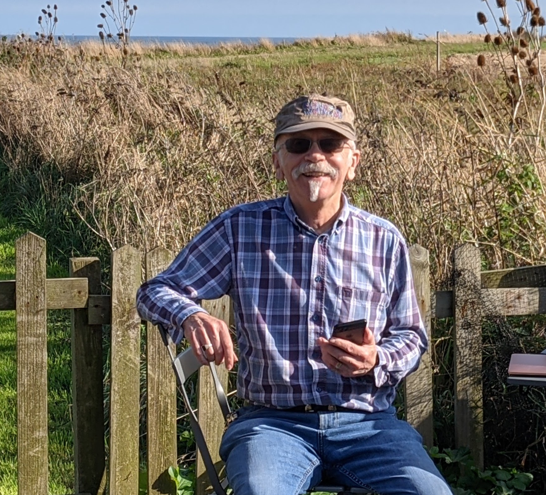 A photo of Steve sitting in a garden. There is a low wooden fence behind him and a field beyond that. You can see the sea in the background..